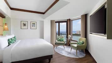 A hotel room with a large bed, two chairs, a TV, and a view of the sea.