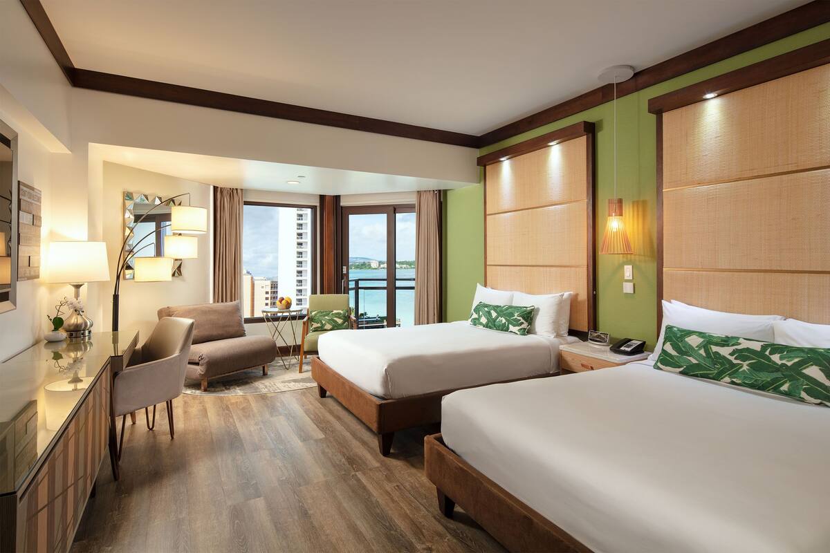 A hotel room with two beds, a desk with a lamp, a chair, and a view of the sea.