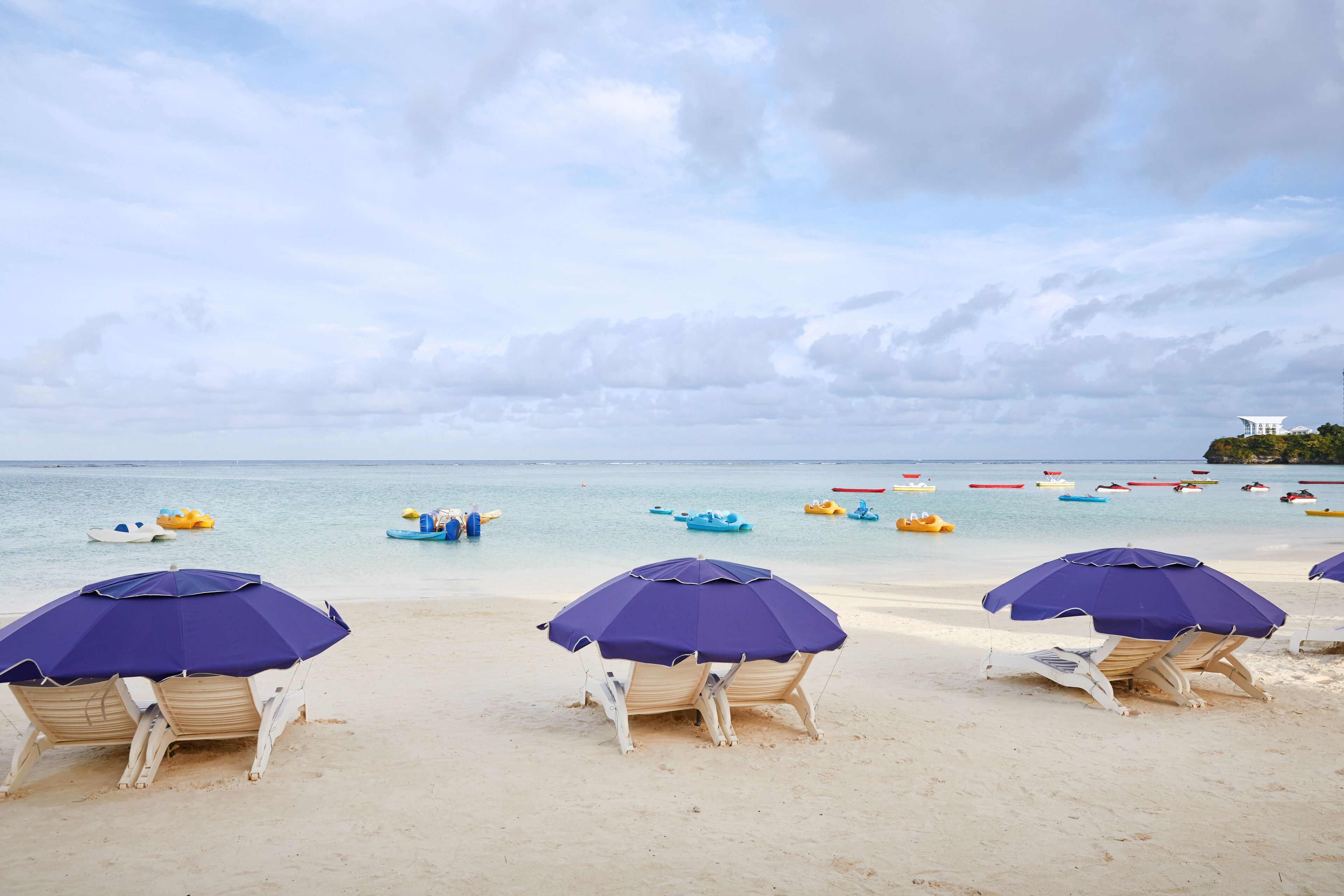 A beach with several purple umbrellas and white lounge chairs, a calm sea, and numerous small boats in the water.