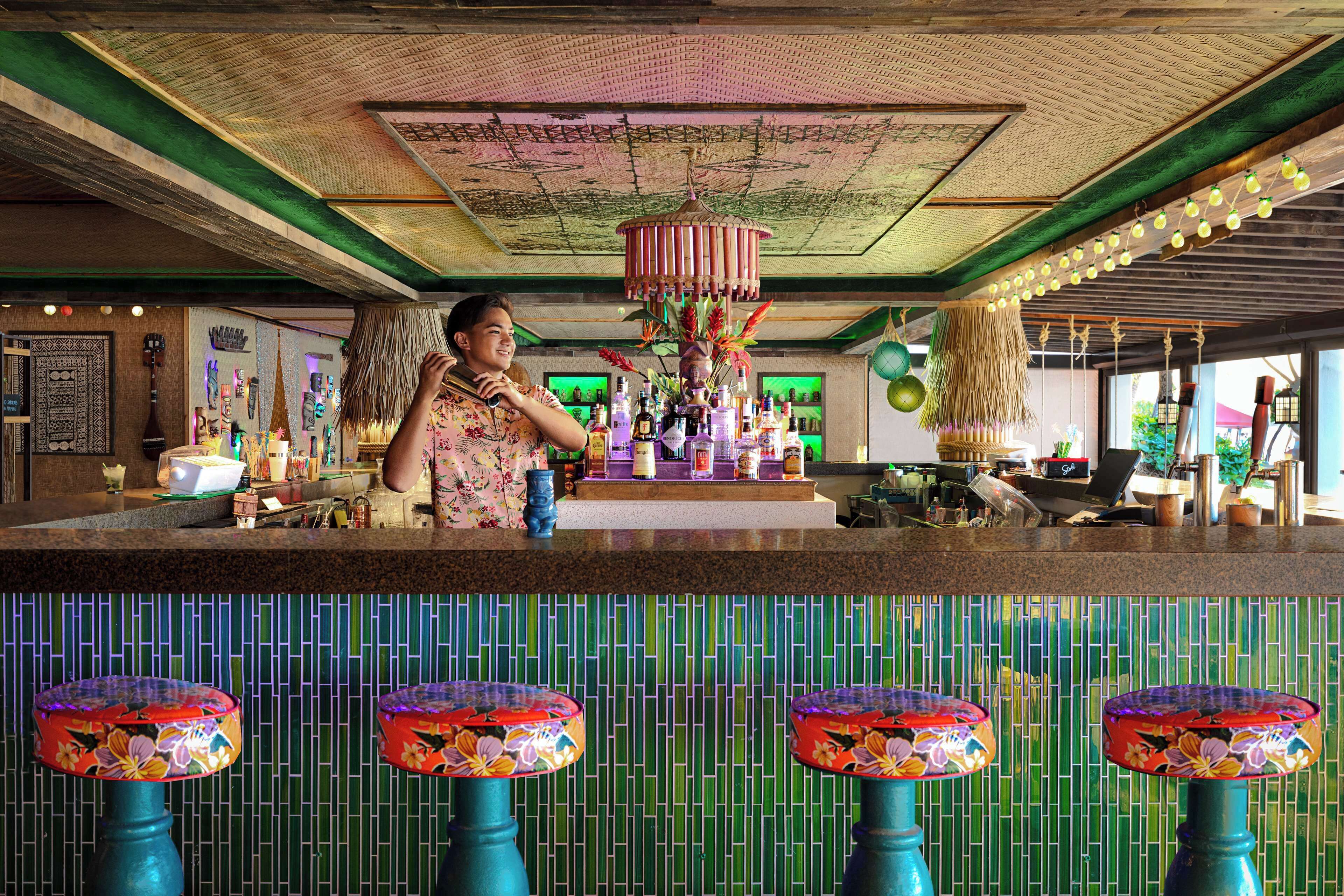 A bar area with a bartender, a decorative hanging light fixture, and a tiled wall with a green and blue pattern.