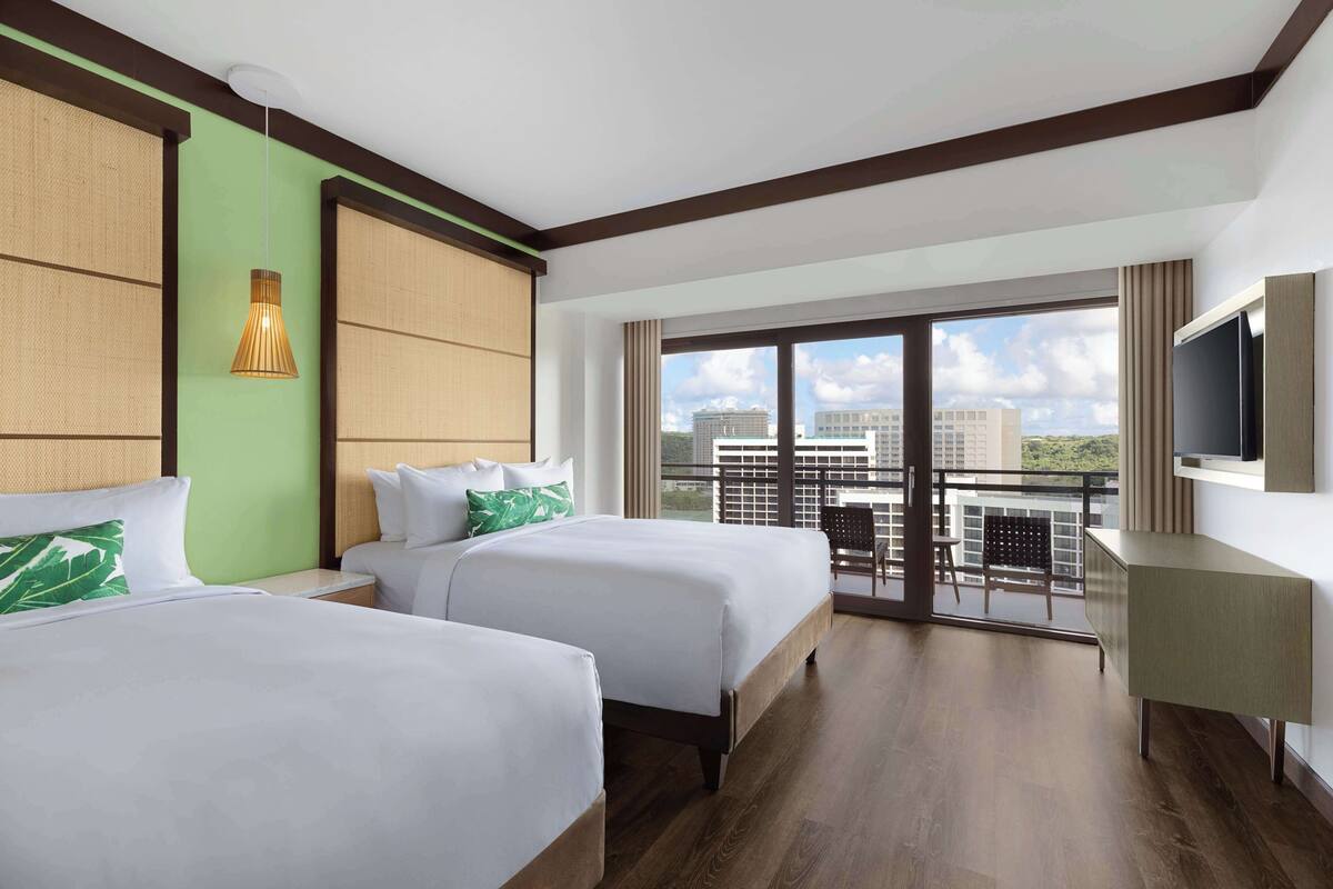 A hotel room with two beds, a balcony with a view of the city, and a flat-screen TV on the wall.