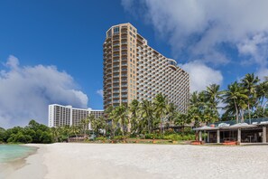 A beachfront hotel with multiple floors, a sandy beach, palm trees, and a clear blue sky. - Dusit Beach Resort Guam (Tamuning)