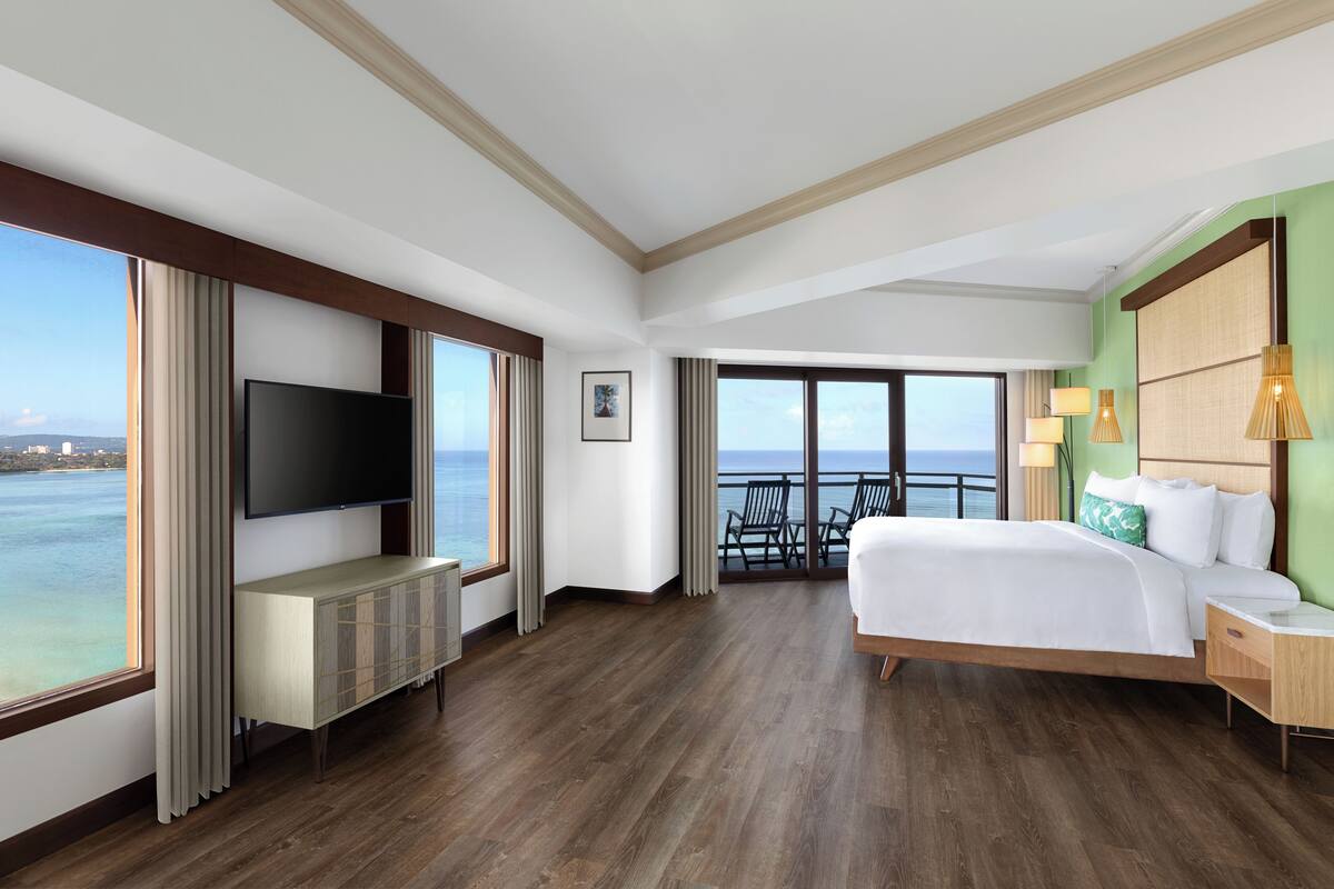 A hotel room with a large TV, a bed, a desk, and a view of the ocean.