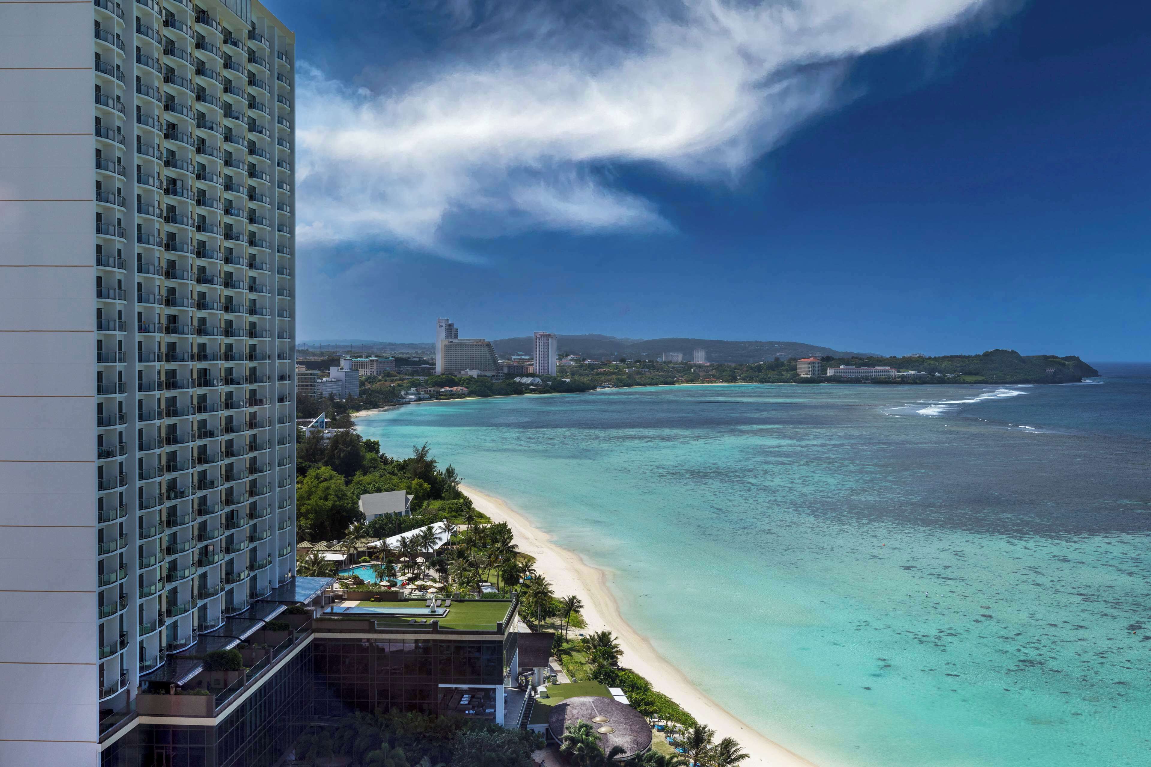 A high-rise hotel overlooking a beach with clear turquoise waters and a distant mountain range.