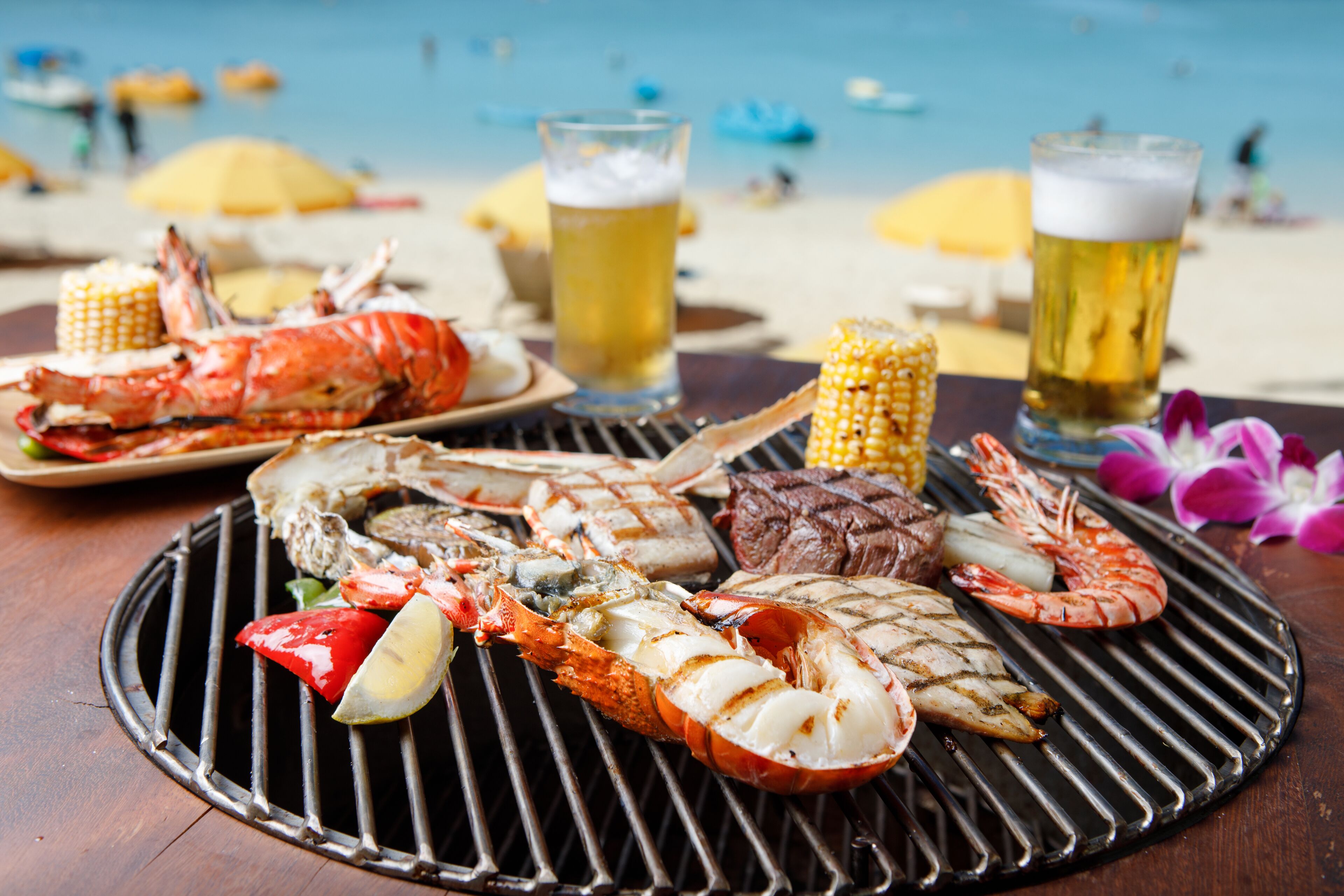 A seafood grill with lobster, shrimp, steak, and corn on a wooden table at a beachside location.