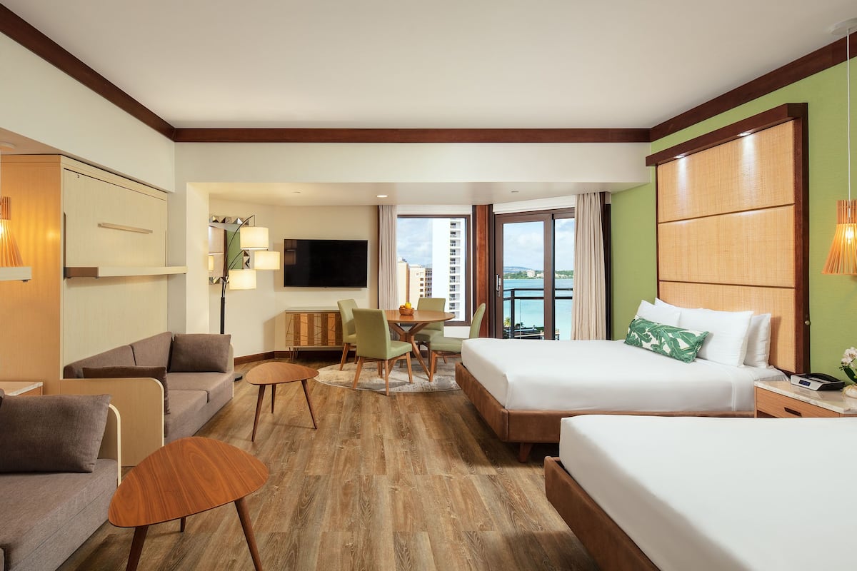 A hotel room with a large bed, a sofa, a dining table with chairs, a TV, and a view of the city.