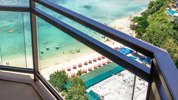 A beach view from a balcony with clear turquoise waters, a sandy beach, and a hotel building in the background. - Dusit Beach Resort Guam (Tamuning)