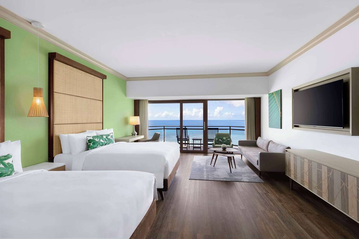 A hotel room with two beds, a sofa, a TV, and a balcony with a view of the sea.