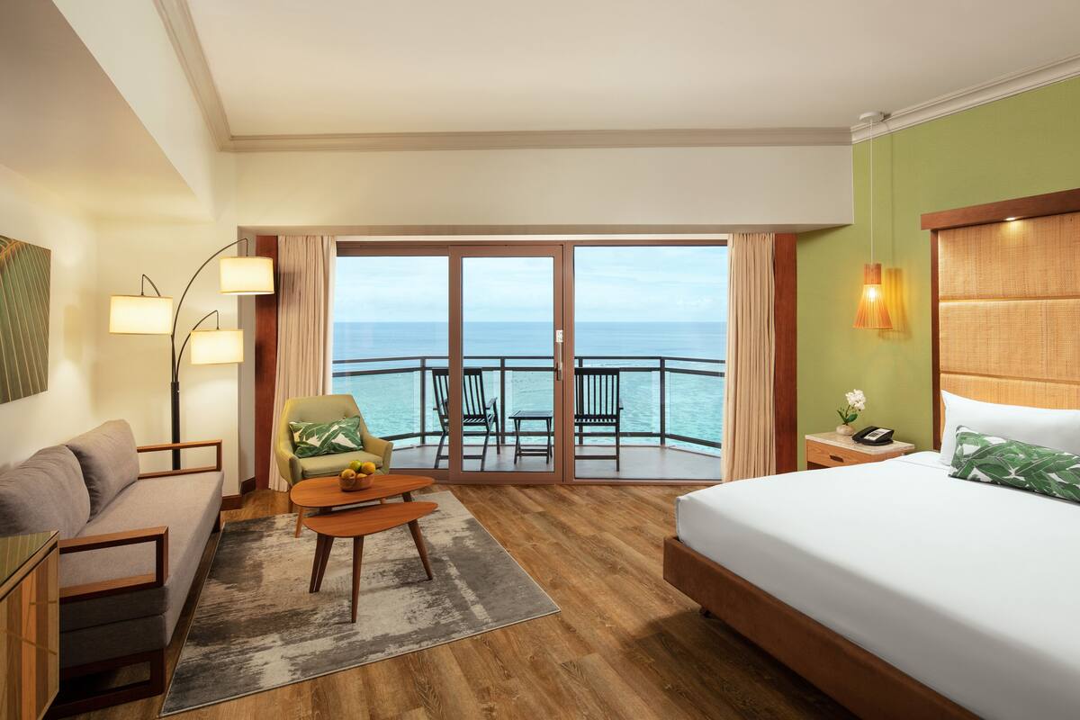 A hotel room with a bed, a sofa, a small table, a chair, a nightstand, and a view of the ocean.