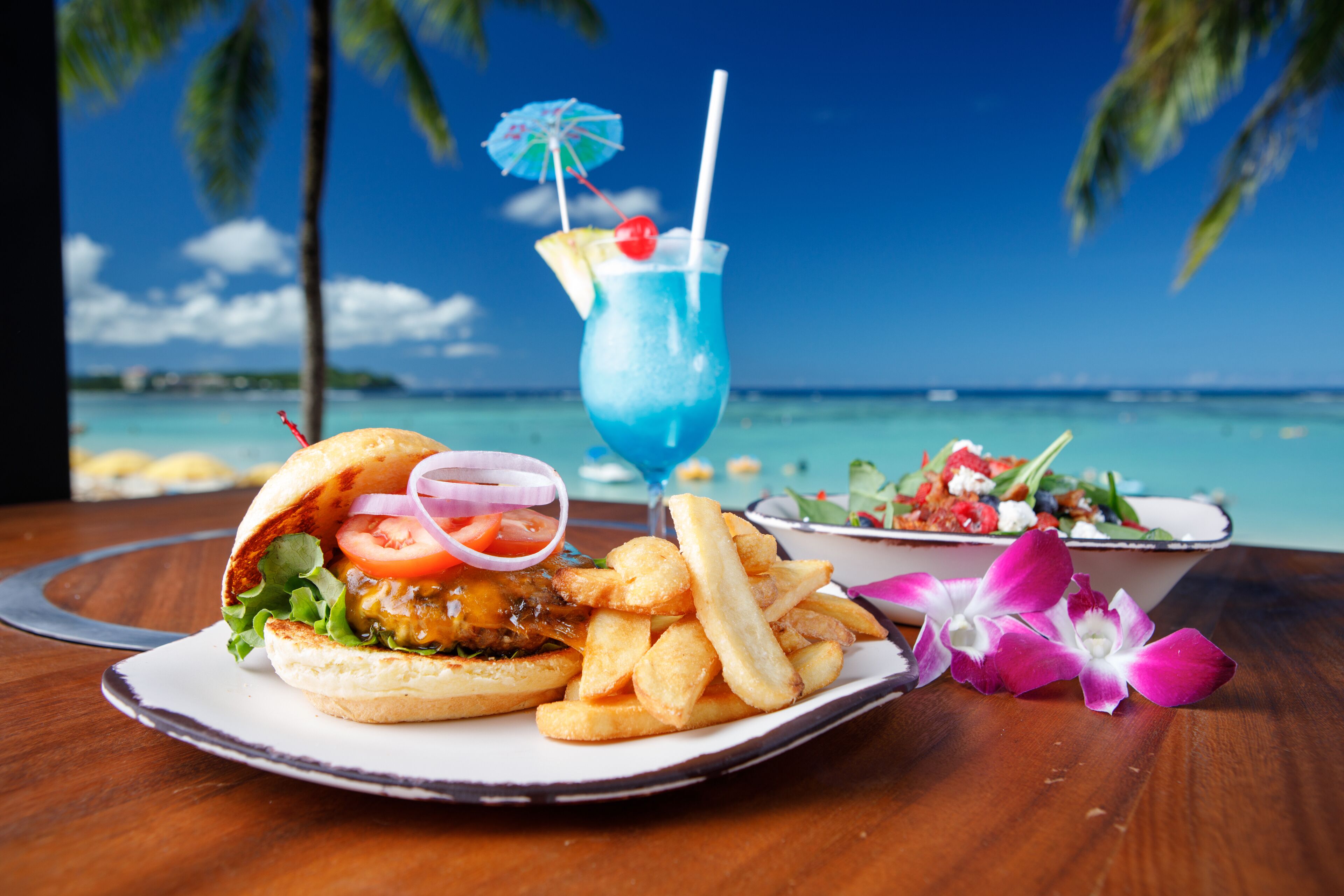 A meal with a burger, fries, and a blue drink on a wooden table with a view of the ocean and palm trees.