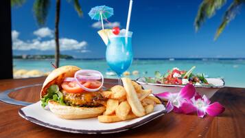 A meal with a burger, fries, and a blue drink on a wooden table with a view of the ocean and palm trees.