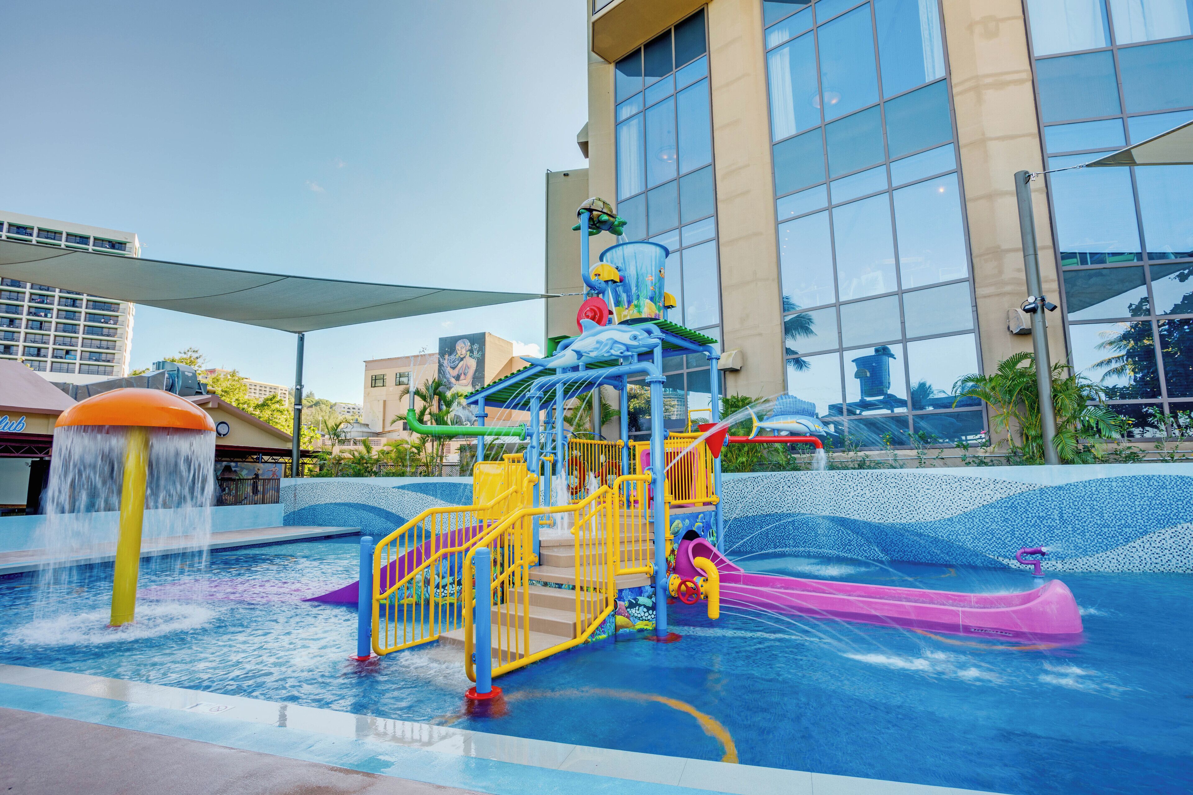 A water play area with slides and water jets, surrounded by a building with large windows.