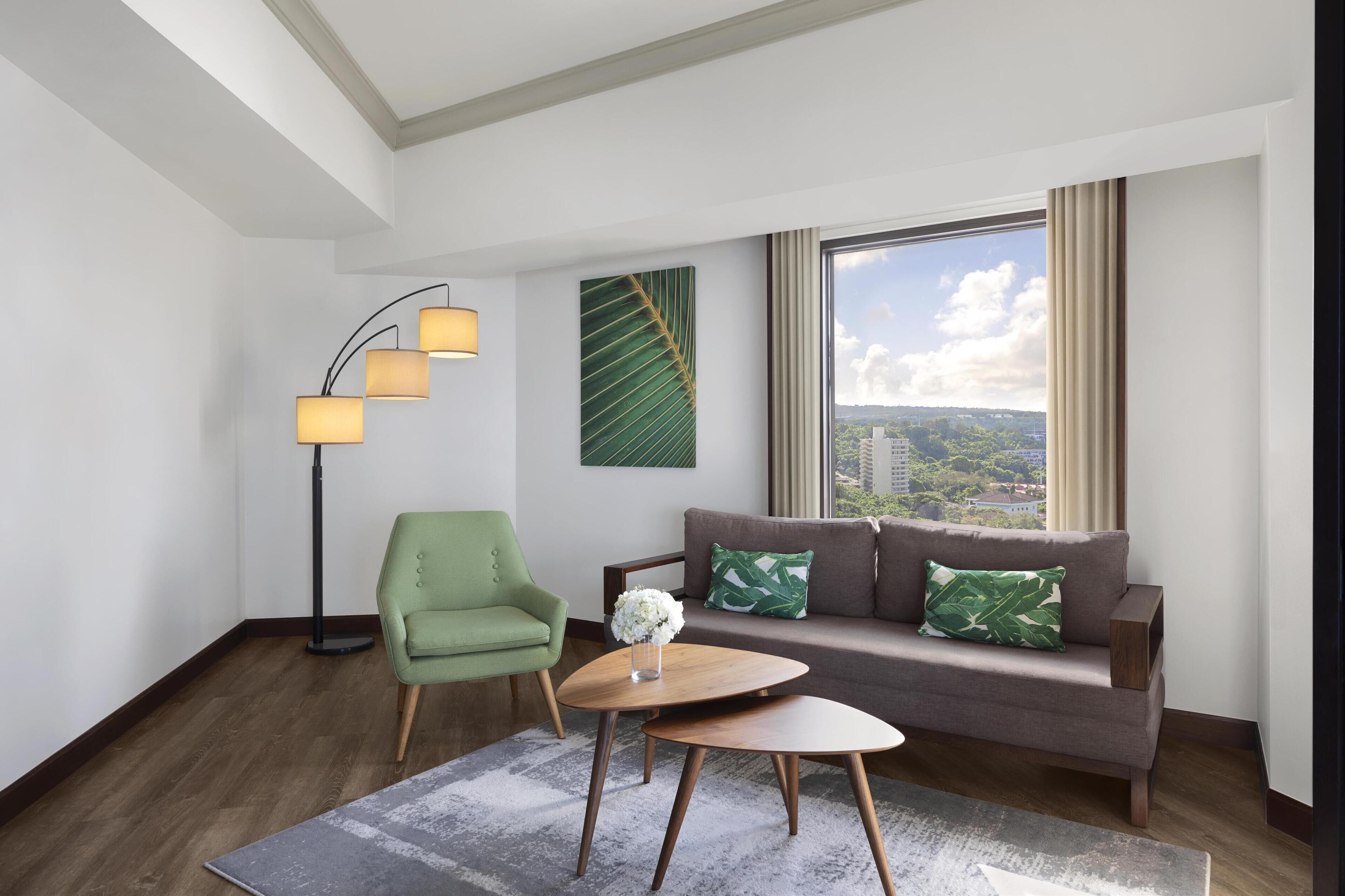 A modern living room with a sofa, a green armchair, a floor lamp, and a view of the city through large windows.