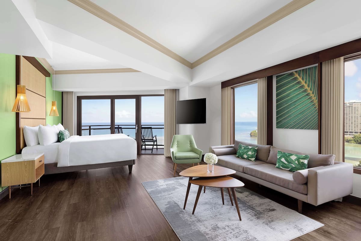 A hotel room with a bed, a sofa, a TV, a chair, a table, and a large window with a view of the sea.