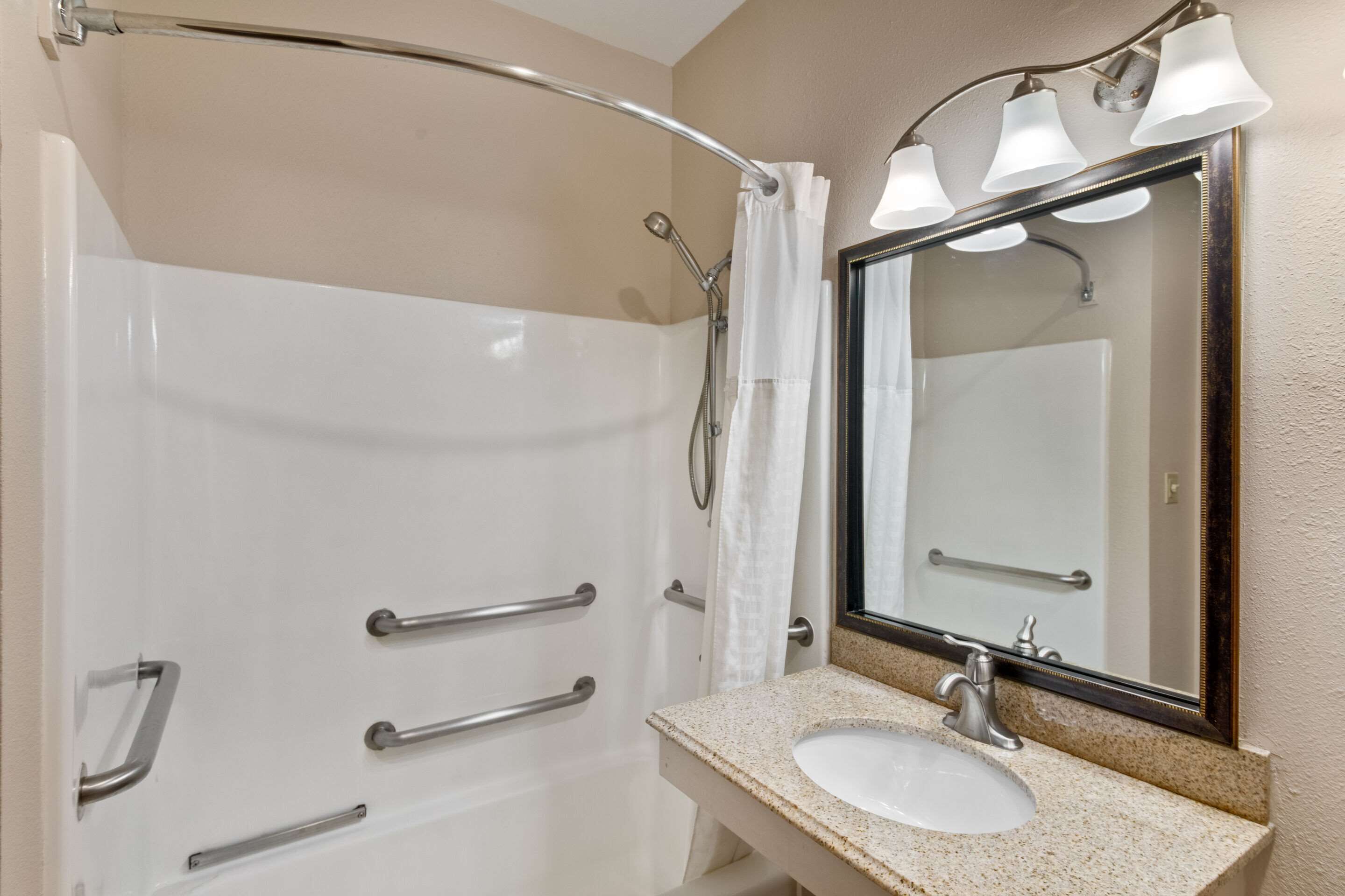 A bathroom with a white bathtub, a sink with a granite countertop, and a mirror with a light fixture above it.