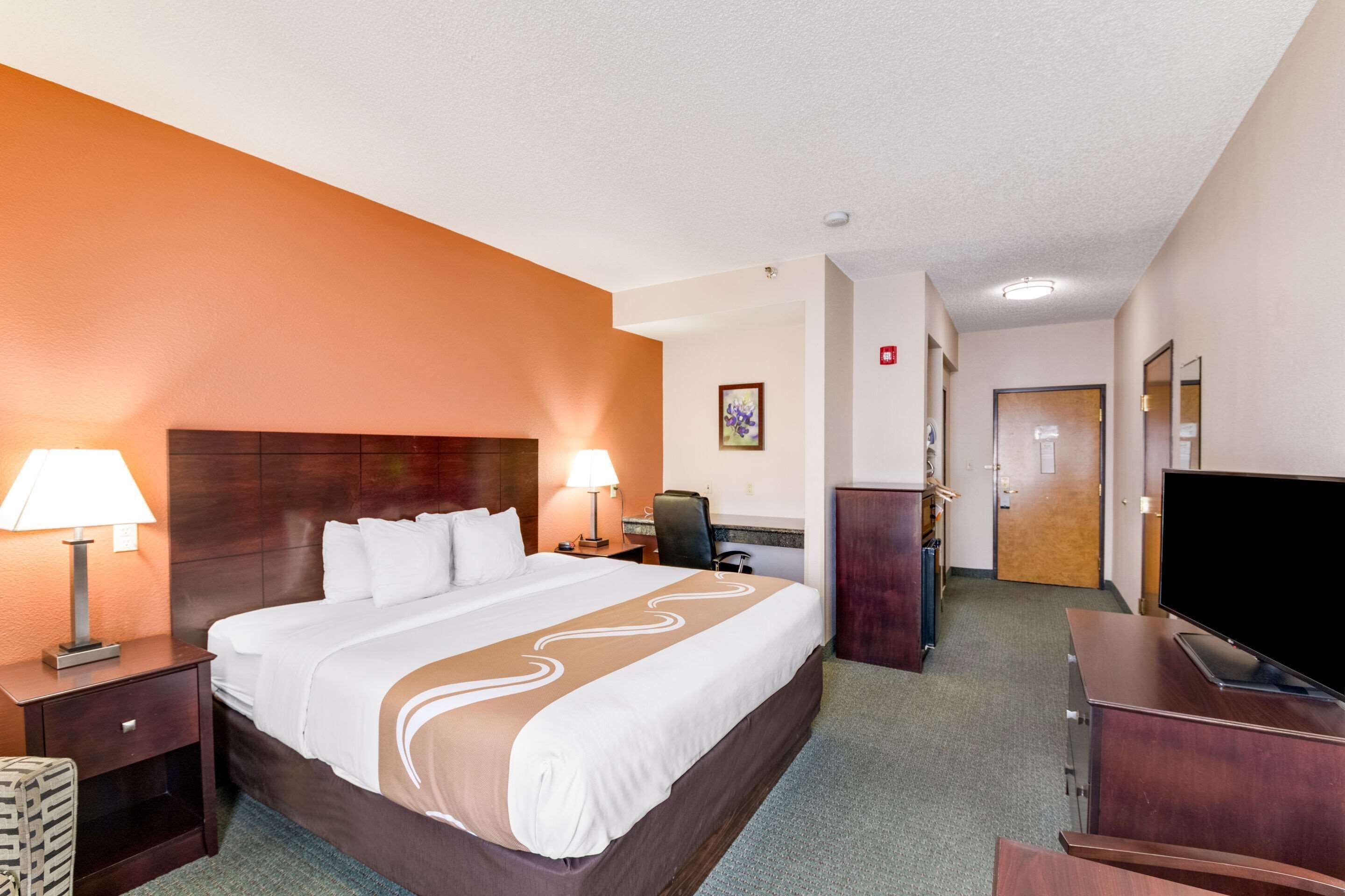 A hotel room with a large bed, two bedside tables with lamps, a desk with a flat-screen TV, a chair, and a wardrobe.
