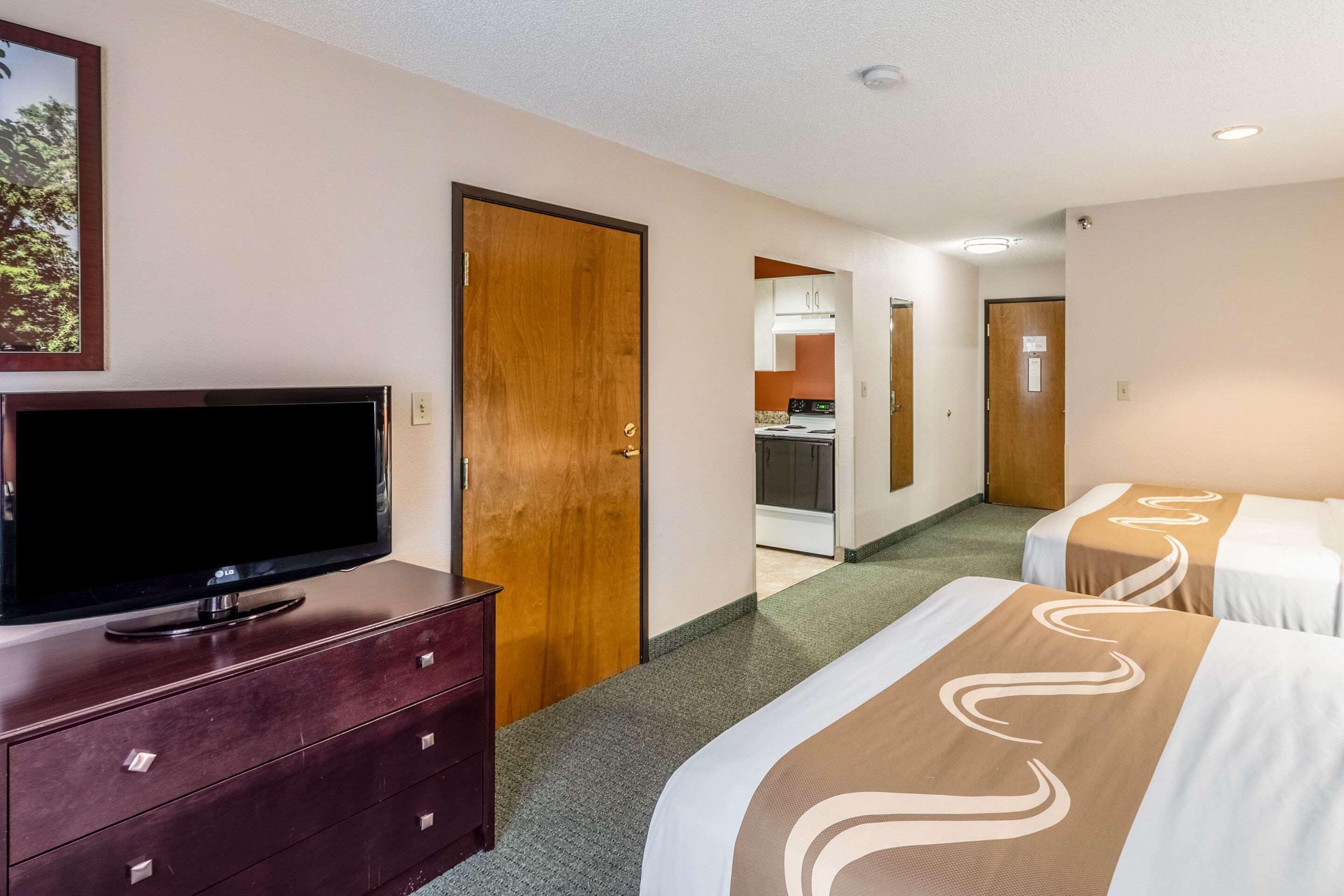A hotel room with a TV on a dresser, two beds with matching headboards, and a hallway leading to a kitchen area.