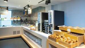 Monipuolinen buffet, jossa on tarjolla erilaisia ruokia ja juomia, kuten hedelmiä, leivonnaisia ja kahviautomaatteja.