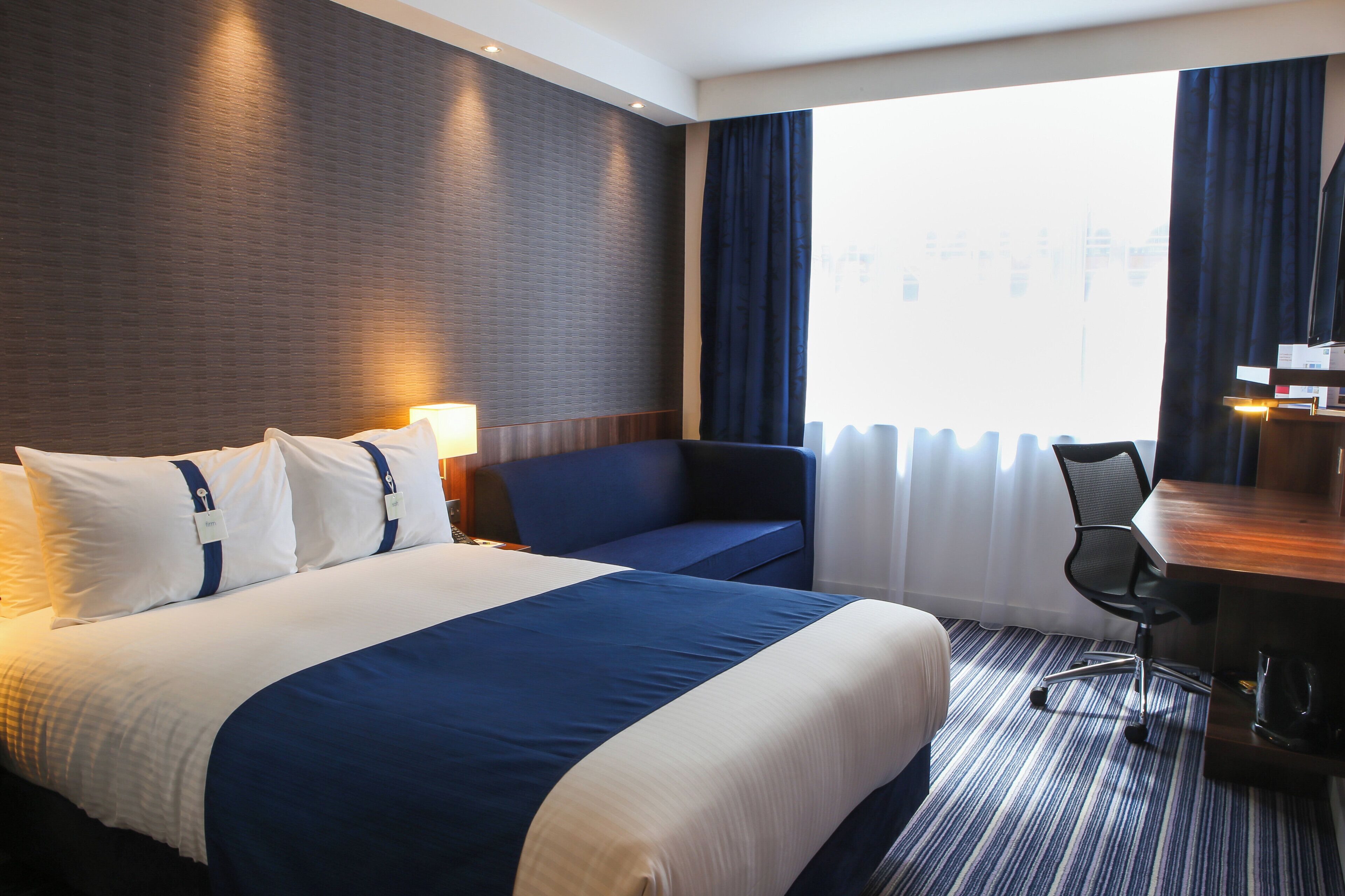 A hotel room with a large bed, a desk, a chair, and a view of the city through the window.