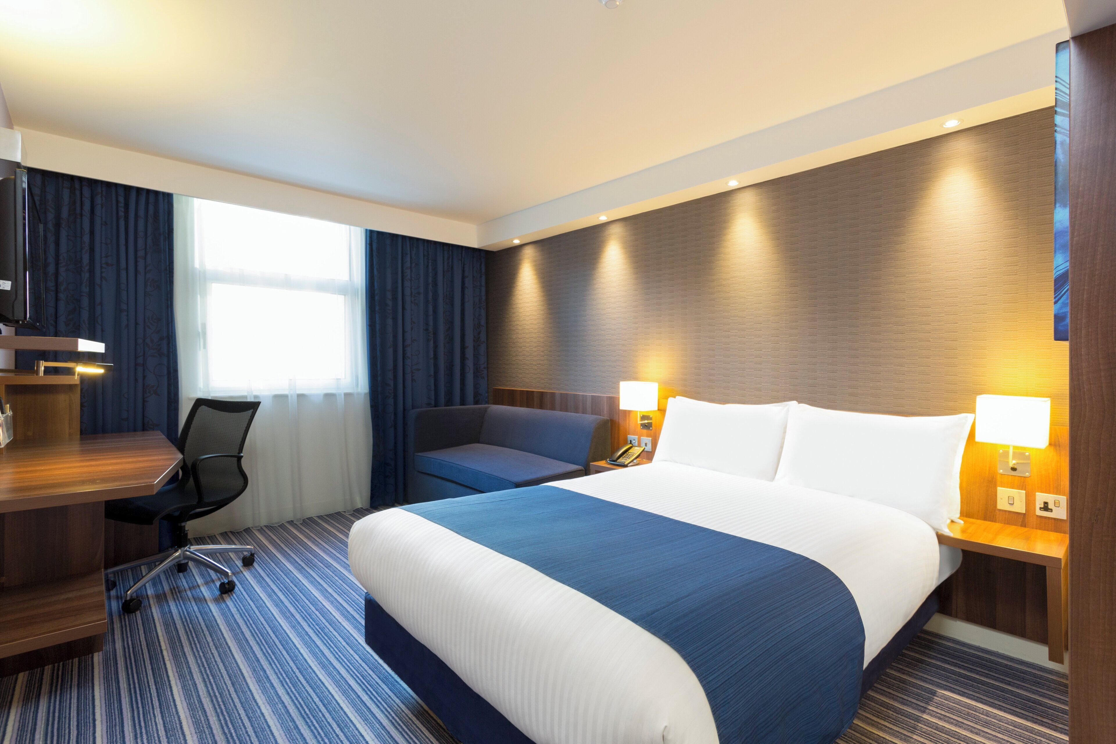 A hotel room with a large bed, a desk, a chair, and a window with curtains.