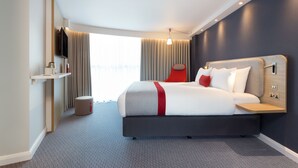 Moderni hotellihuone, jossa on suuri sänky, seinälle asennettu taulutelevisio, pieni pöytä ja ikkuna, jossa on verhot.