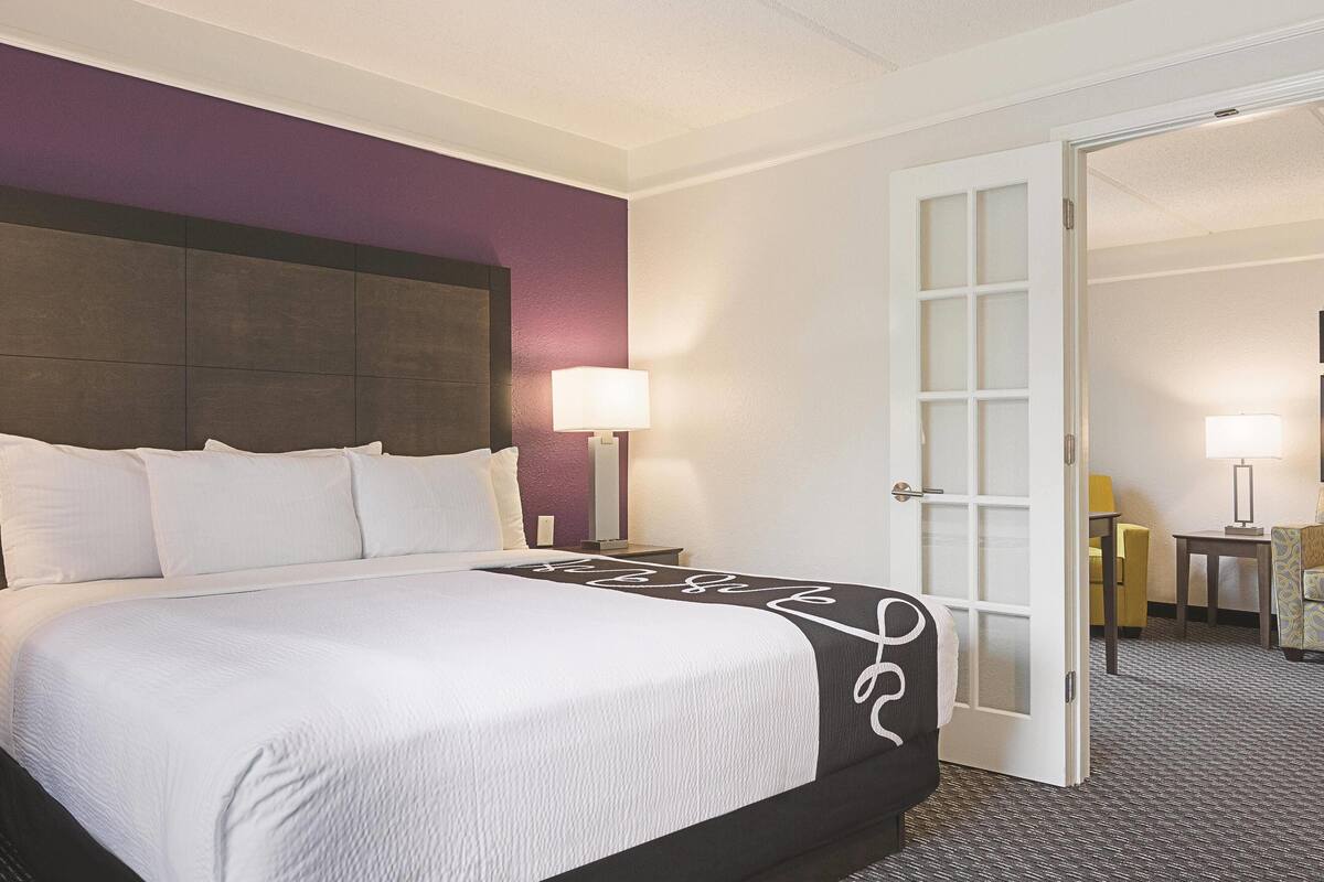 A hotel room with a bed, a nightstand, a lamp, and a door leading to another room.
