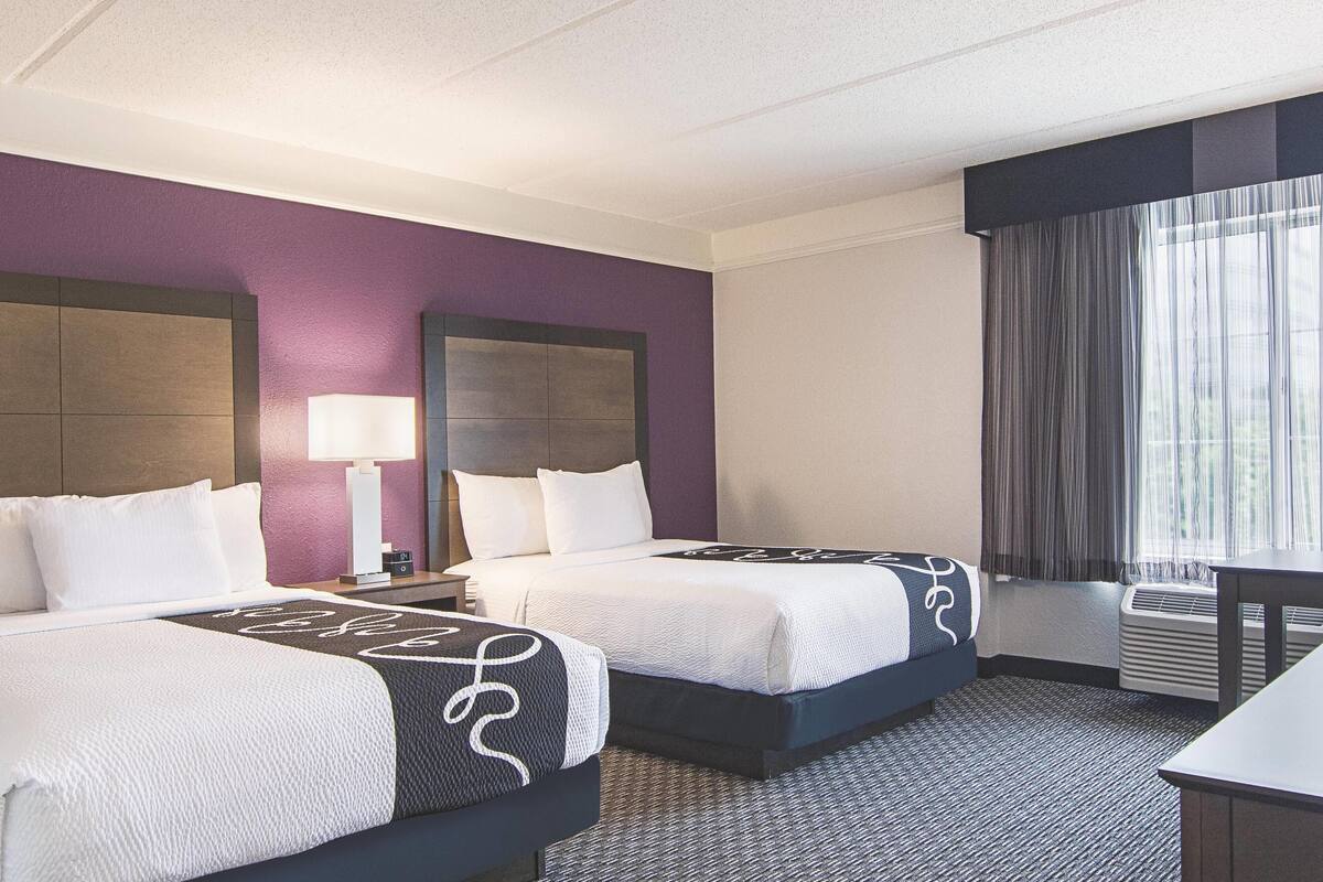 A hotel room with two beds, a desk, a chair, a lamp, and a window with curtains.