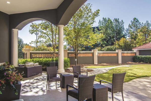 Une terrasse extérieure aménagée avec des meubles en osier, un espace couvert et une vue sur un jardin paysager et des arbres.