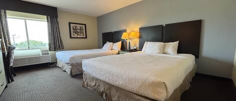 A hotel room with two beds, a window with curtains, a wall-mounted picture, and a lamp.