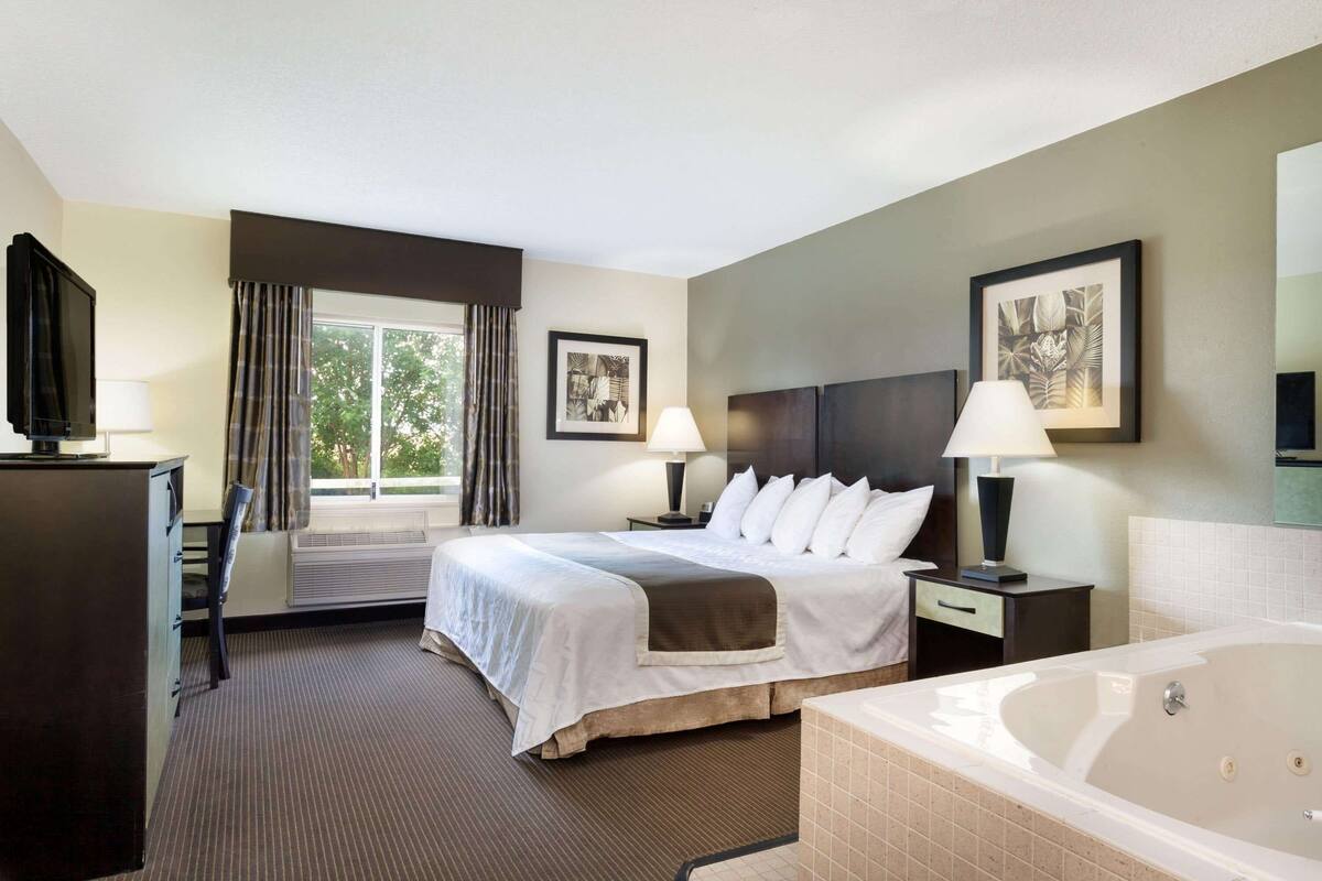 A hotel room with a bed, bedside lamps, a desk, a television, a window with curtains, a framed picture, and a bathtub.