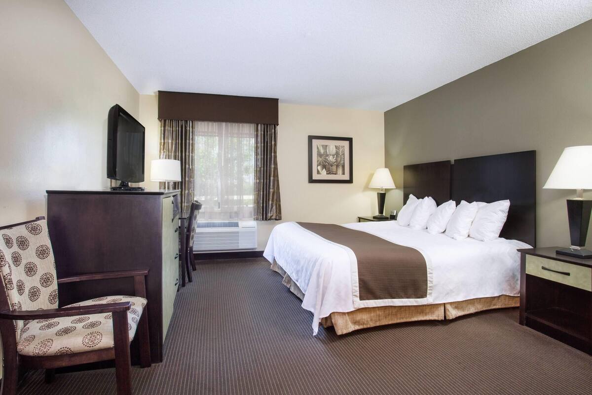 A hotel room with a bed, a desk with a TV, a chair, a window with curtains, and a picture on the wall.