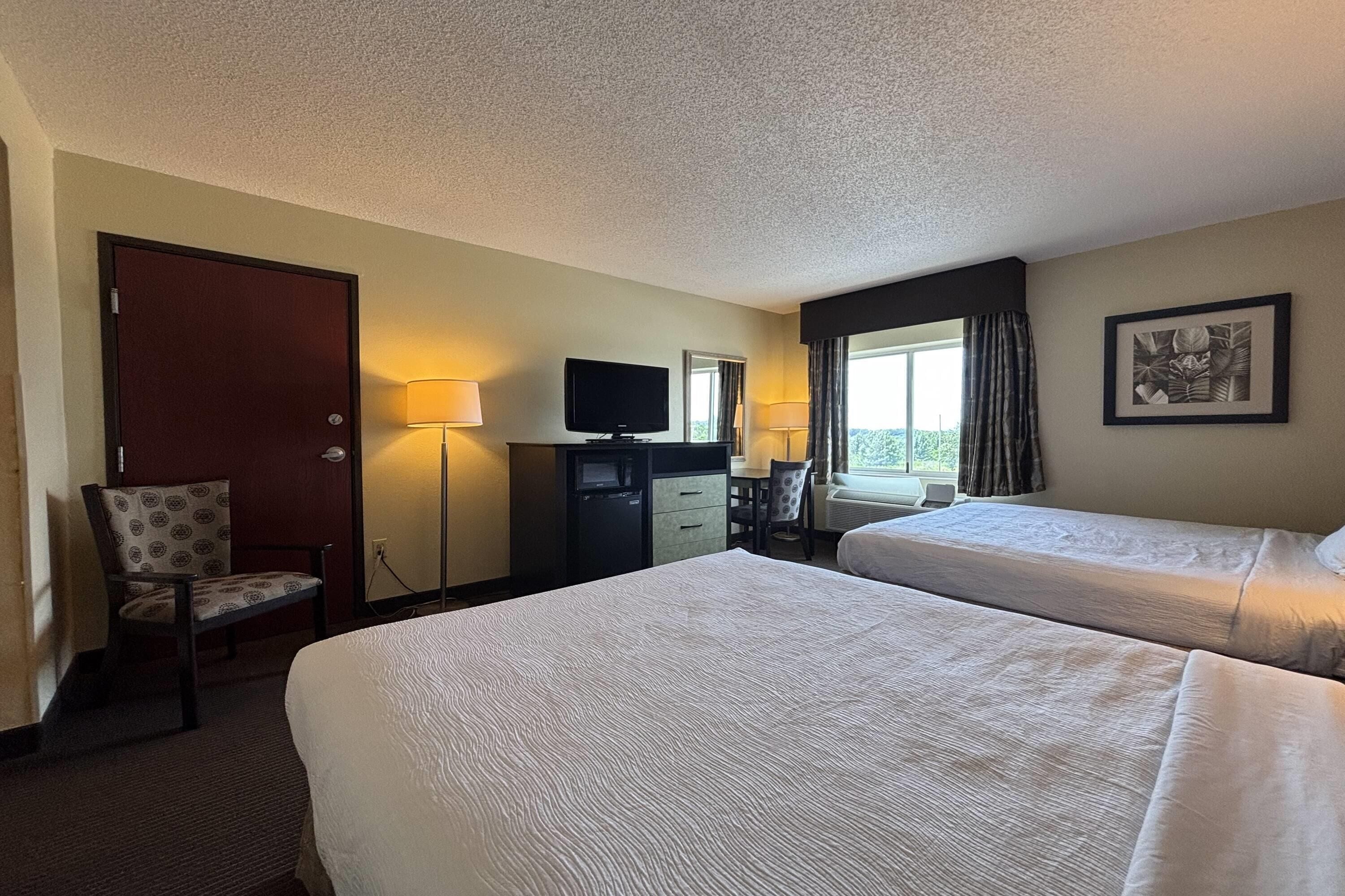 A hotel room with two beds, a desk with a TV, a chair, a desk lamp, and a window with curtains.