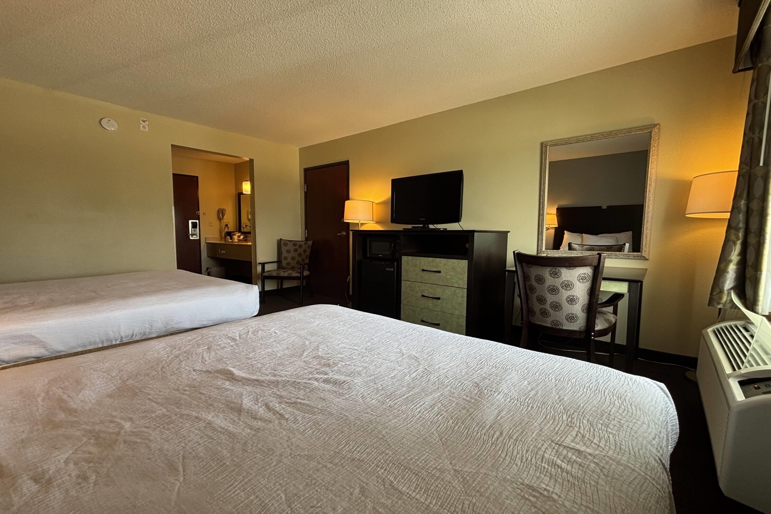A hotel room with two beds, a desk with a television, a chair, a mirror, and a heater.