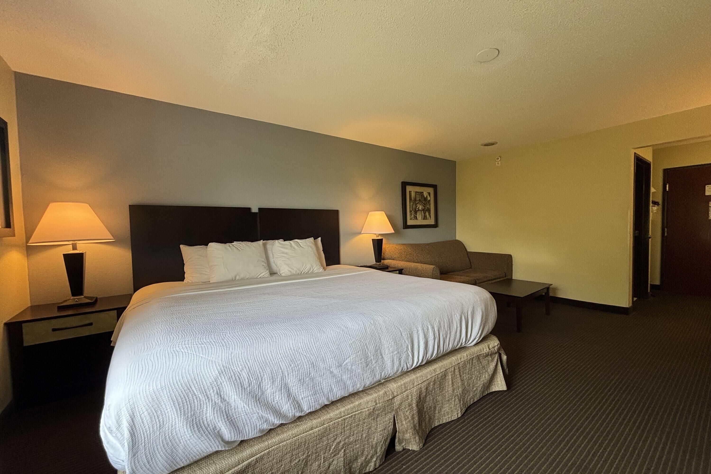 A hotel room with a large bed, two bedside tables with lamps, a nightstand, a sofa, and a door.