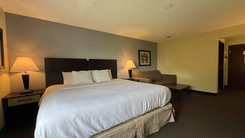 A hotel room with a large bed, two bedside tables with lamps, a nightstand, a sofa, and a door.