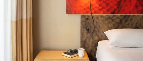 Une chambre d’hôtel avec un lit, une table de chevet sur laquelle se trouvent un livre et une tasse, une moquette rouge et une lampe fixée au mur.