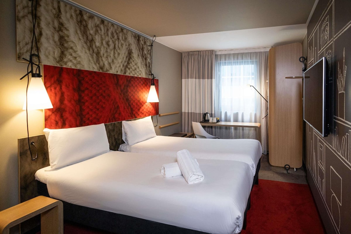 Ett hotellrum med två sängar, ett skrivbord, en stol, en TV och ett fönster med gardiner.