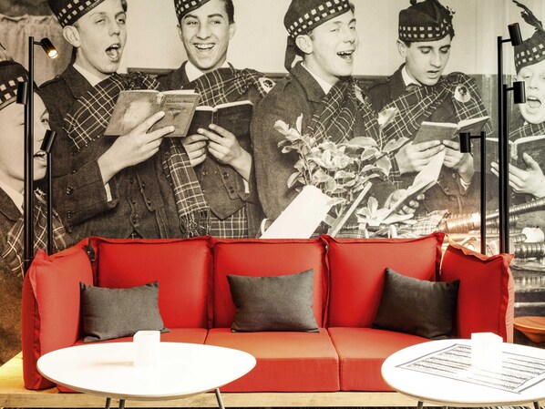 A red sofa with grey cushions, a white table, and a wall with a black and white photo. - ibis Edinburgh Centre Royal Mile (Edinburgh)