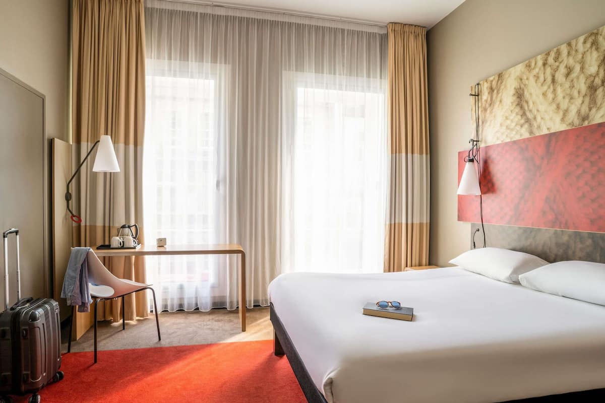 Une chambre d’hôtel avec un lit, un bureau doté d’une lampe, une valise et une vue sur la ville grâce à de grandes fenêtres.