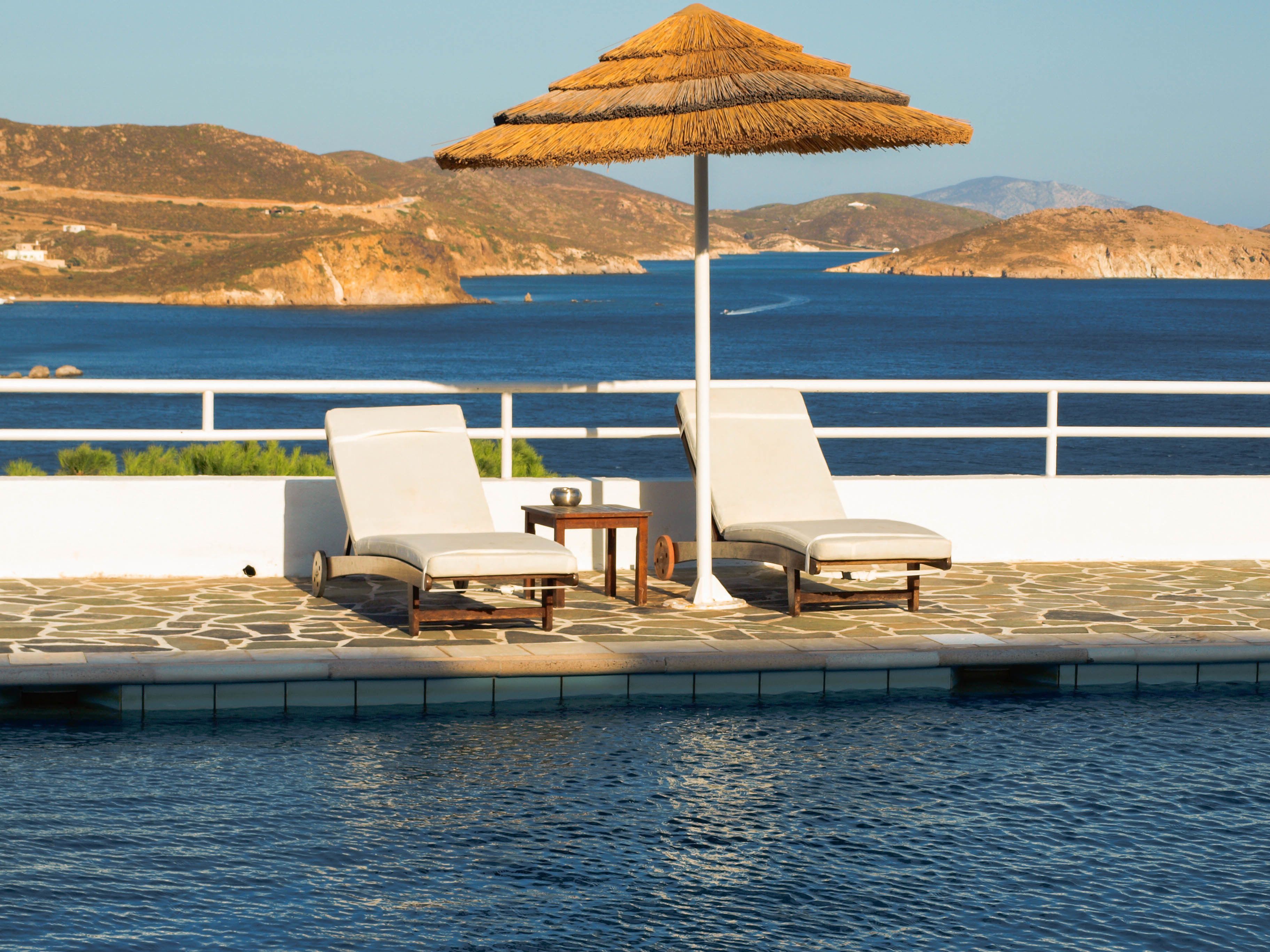 A poolside area with two lounge chairs and a table, a thatched umbrella, and a view of the sea and hills.