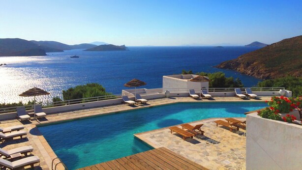 A pool with clear blue water, surrounded by lounge chairs and umbrellas, overlooking a coastal landscape with mountains and the sea.