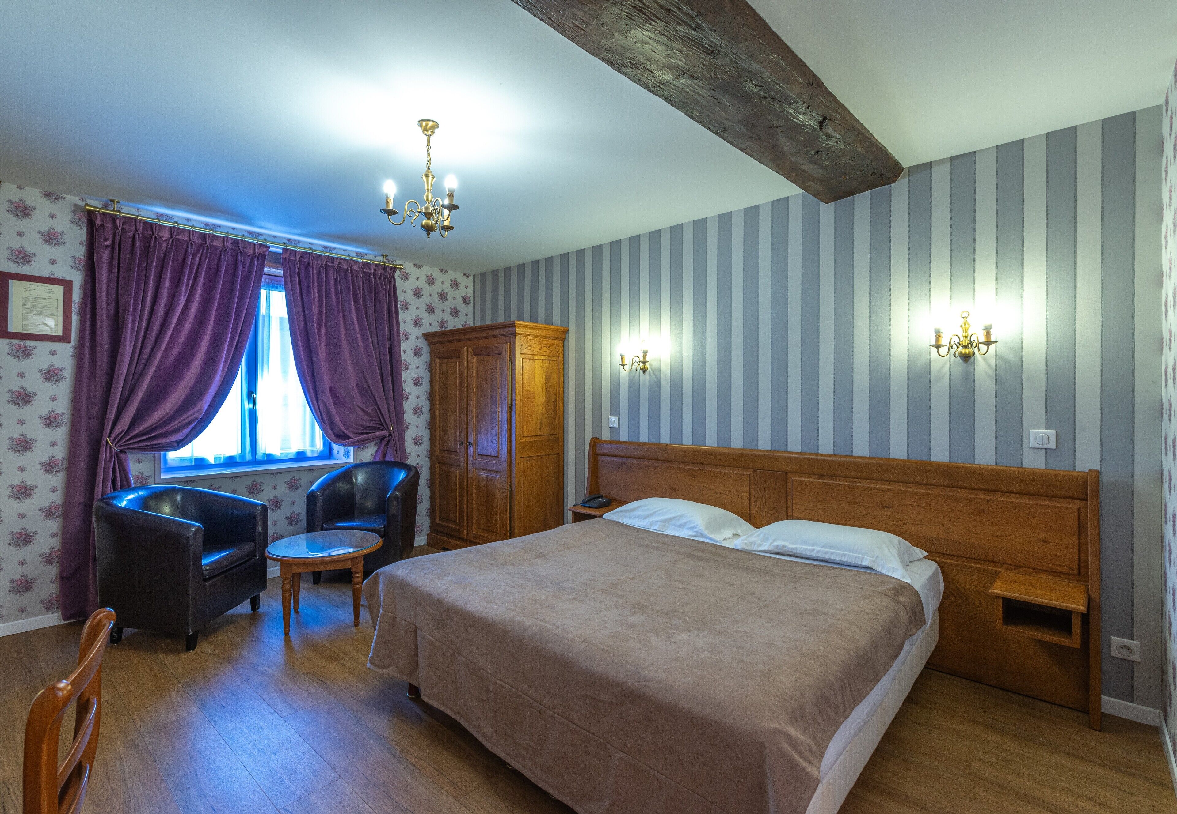 Een slaapkamer met een bed, twee stoelen, een houten garderobe, een klein tafeltje en een raam met gordijnen.