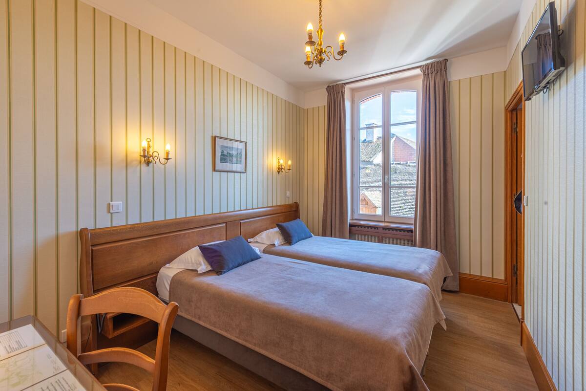 Une chambre d’hôtel avec deux lits, des têtes de lit en bois, un papier peint à rayures, un lustre, une fenêtre avec des rideaux et un téléviseur fixé au mur.