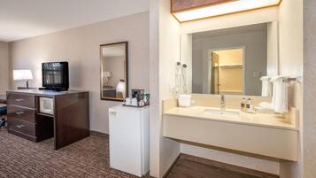 Quarto de hotel com escrivaninha, televisão, minibar e banheiro com pia, espelho e porta-toalhas.