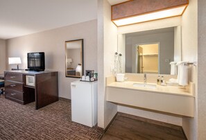 Une chambre d’hôtel équipée d’un bureau, d’une télévision, d’un minibar et d’une salle de bain avec un lavabo, un miroir et un porte-serviettes.