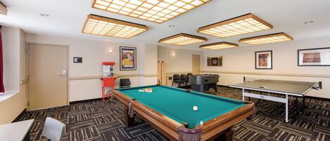 A pool table with a green felt surface, a foosball table, and a dartboard in a room with patterned carpet and recessed ceiling lighting.