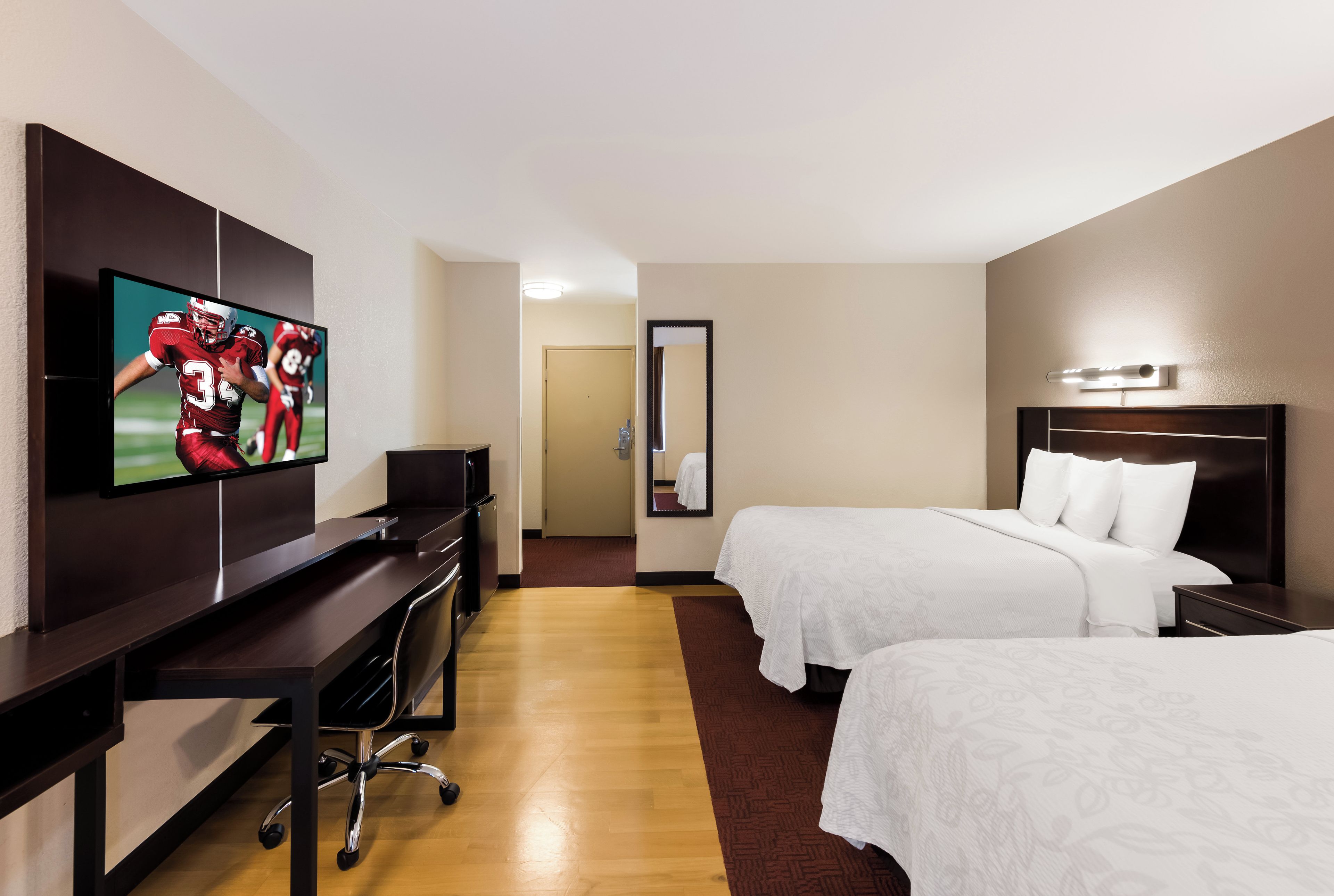 A hotel room with a flat-screen TV, a desk, two beds, a mirror, and a door leading to another room.