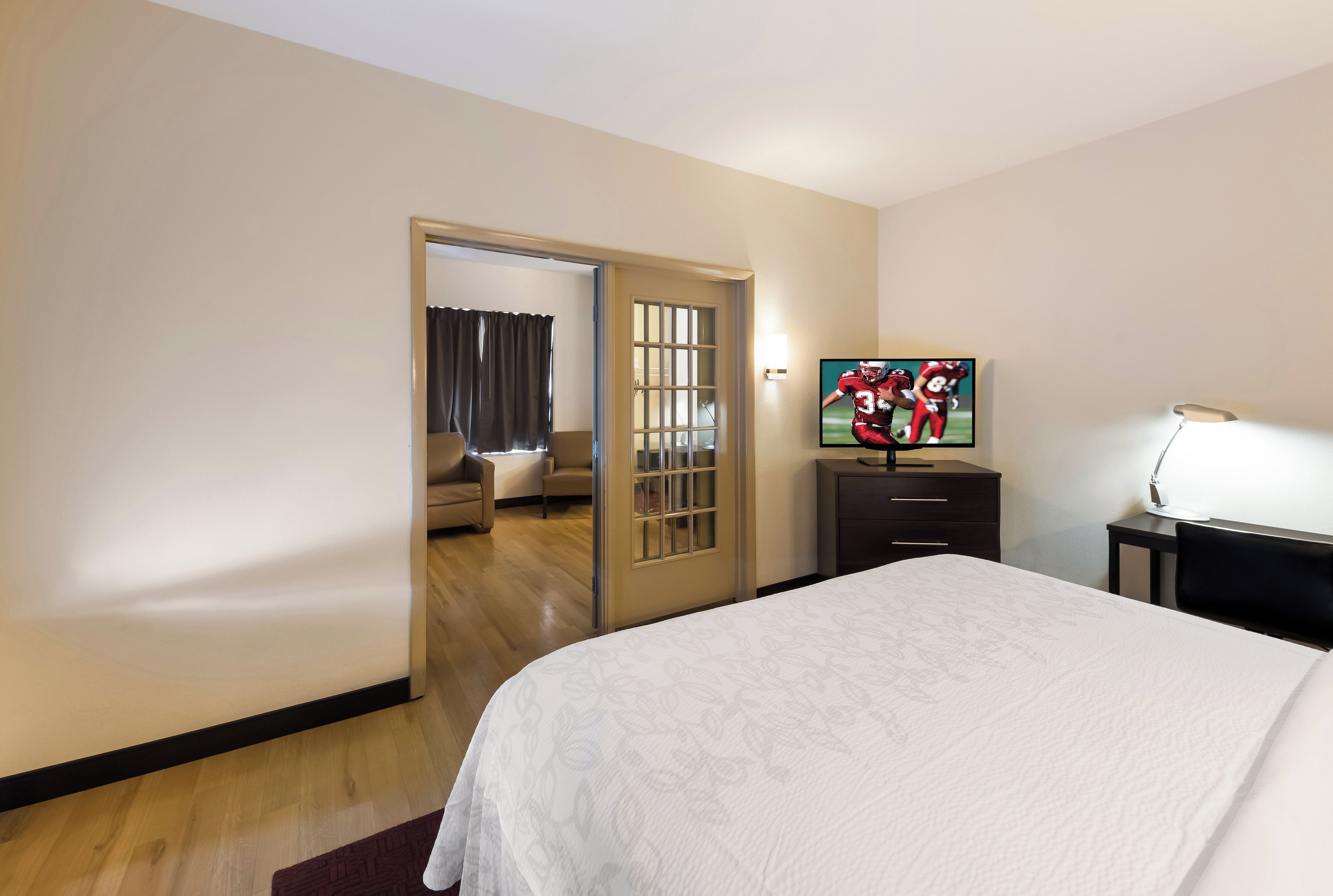 A hotel room with a bed, a television, a desk with a lamp, and a door leading to a sitting area.