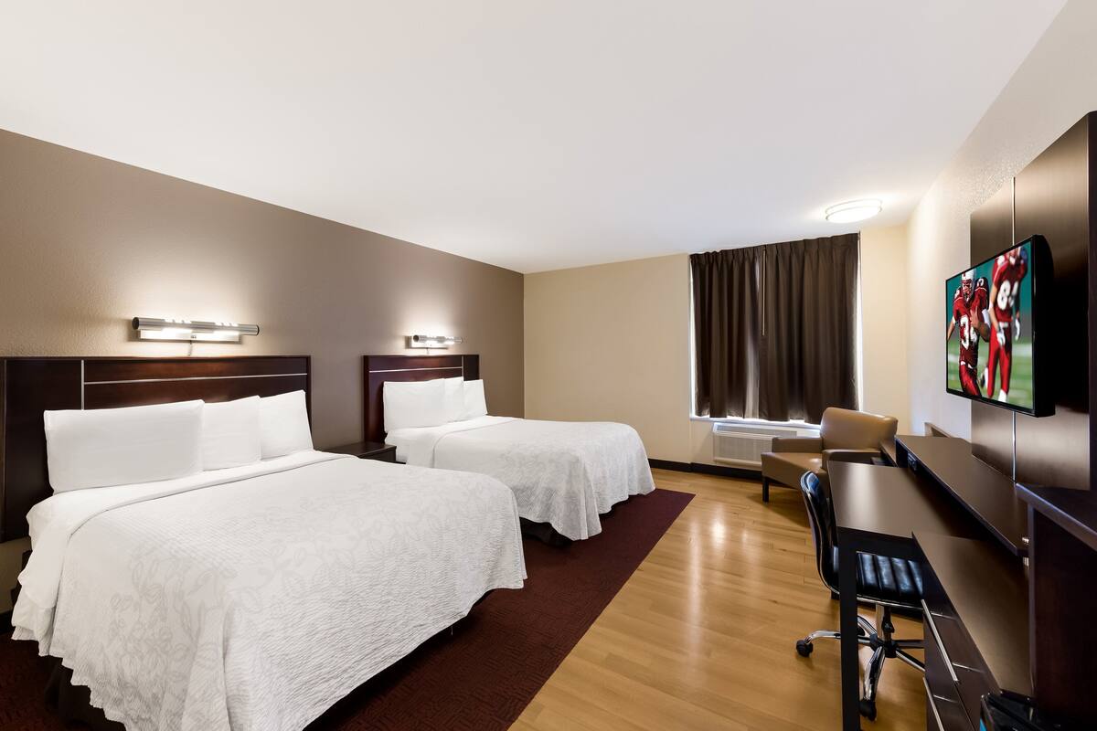 A hotel room with two beds, a desk, a chair, a TV, and a window with curtains.