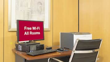 Habitación de hotel con escritorio, computadora, impresora y una silla. Un cartel en el escritorio anuncia Wi-Fi gratuito en todas las habitaciones.